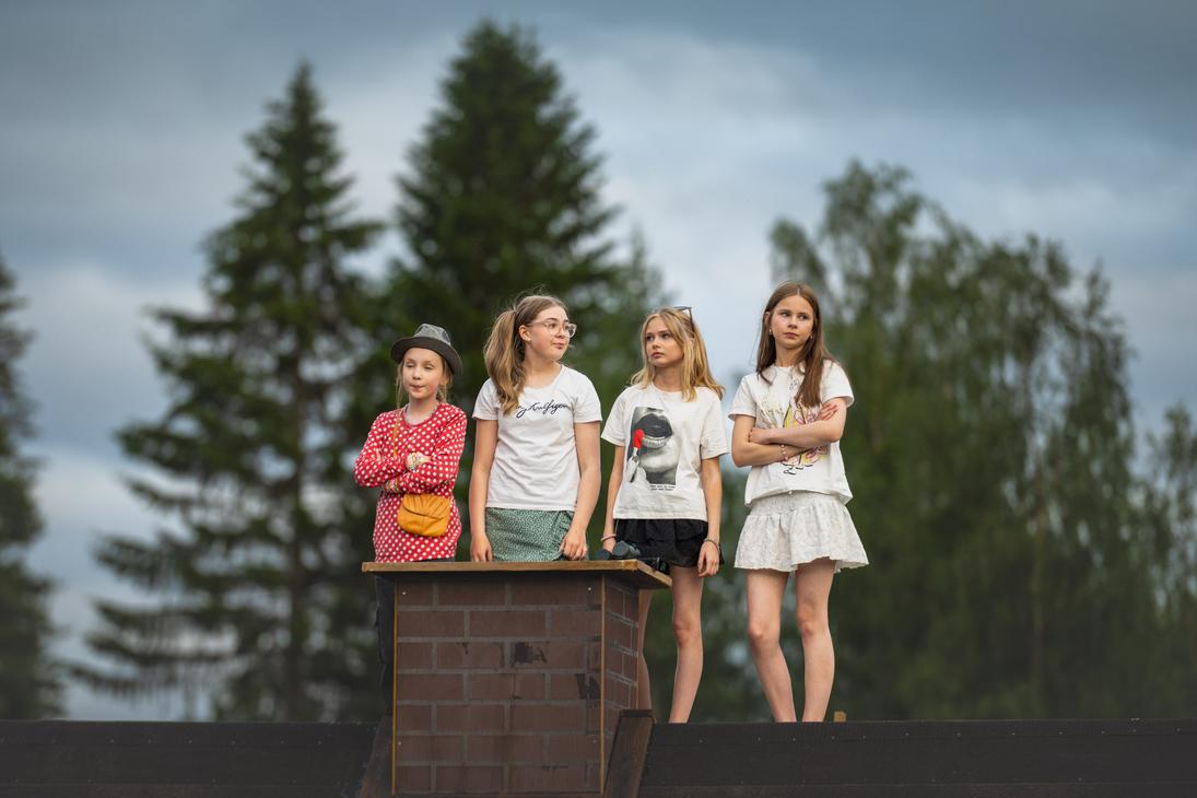 Neljä nuorta tyttöä seisoo vierekkäin tiilisavupiipun vieressä rakennuksen katolla Möhkön kesäteatterin Peitetarina-näytelmässä. Heillä on yllään kesäiset vaatteet, osa pitää käsiään puuskassa, ja he katsovat tarkkaavaisina sivuun taustallaan korkeat vihreät puut ja pilvinen taivas.