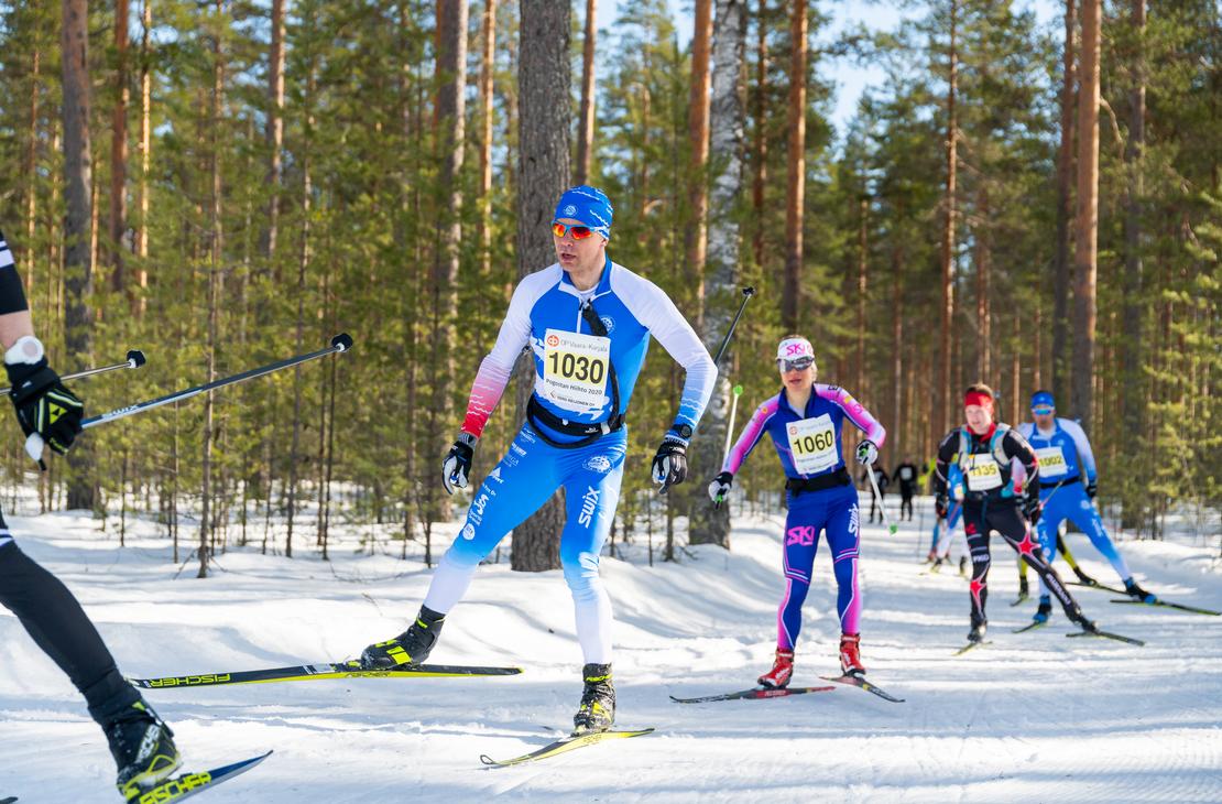 uva hiihtokilpailusta aurinkoisessa, lumisessa mets&auml;maastossa. Etualalla useita hiiht&auml;ji&auml; on liikkeess&auml;. Keskeisen&auml; hahmona on mies, jolla on sinivalkoinen, numerolla 1030 varustettu hiihtoasu, ja h&auml;n k&auml;ytt&auml;&auml; luistelutyyli&auml;. H&auml;nen takanaan on nainen pinkin- ja sinis&auml;vyisess&auml; asussa, numerolla 1060, ja muita hiiht&auml;ji&auml; kauempana. Maasto on tasaista, ja taustalla on tihe&auml;, m&auml;ntyvaltainen mets&auml;.