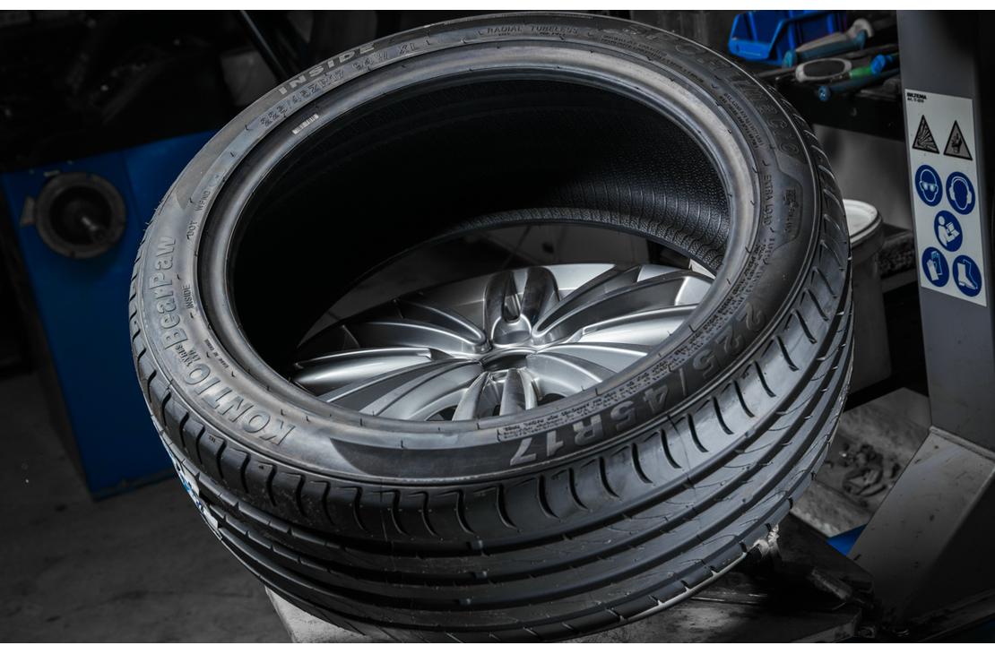 Lähikuva uudesta, matalaprofiilisesta renkaasta, joka on asennettavana kiiltävälle alumiinivanteelle. Renkaan pintakuvio on selkeä ja se on asetettu kaltevassa asennossa todennäköisesti rengaskoneen päällä. Taustalla näkyy epätarkkana sinisiä ja harmaita osia rengashuoltovälineistöä.