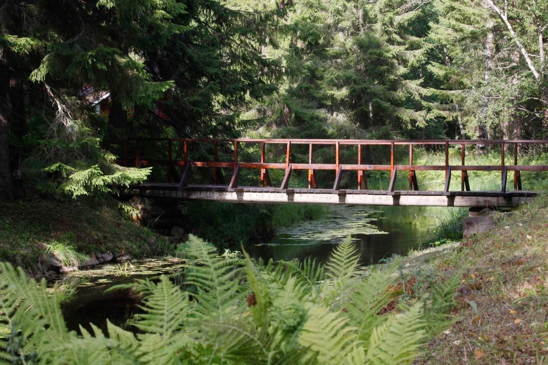Ruskea puinen silta ylittää kapean puron Möhkön ruukkimuseon vehreässä metsämaisemassa. Etualalla kasvaa saniaisia ja veden pinnalla kelluu lumpeenlehtiä.