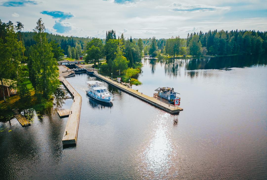 Ilmakuva tyynestä vesistöstä aurinkoisena päivänä, joka liittyy Blue Lake Cruisesin toiminta-alueeseen. Kuvassa näkyy kaksi venelaituria ja niihin kiinnitettynä useita aluksia, mukaan lukien suurempi, valkoinen risteilyalus ja pienempi vene. Laitureiden takana näkyy osa maata, mahdollisesti kanava tai sulku (vasemmalla), ja aluetta ympäröi tiheä, vehreä metsä. Auringonvalo heijastuu kirkkaasti veden pintaan oikealla.
