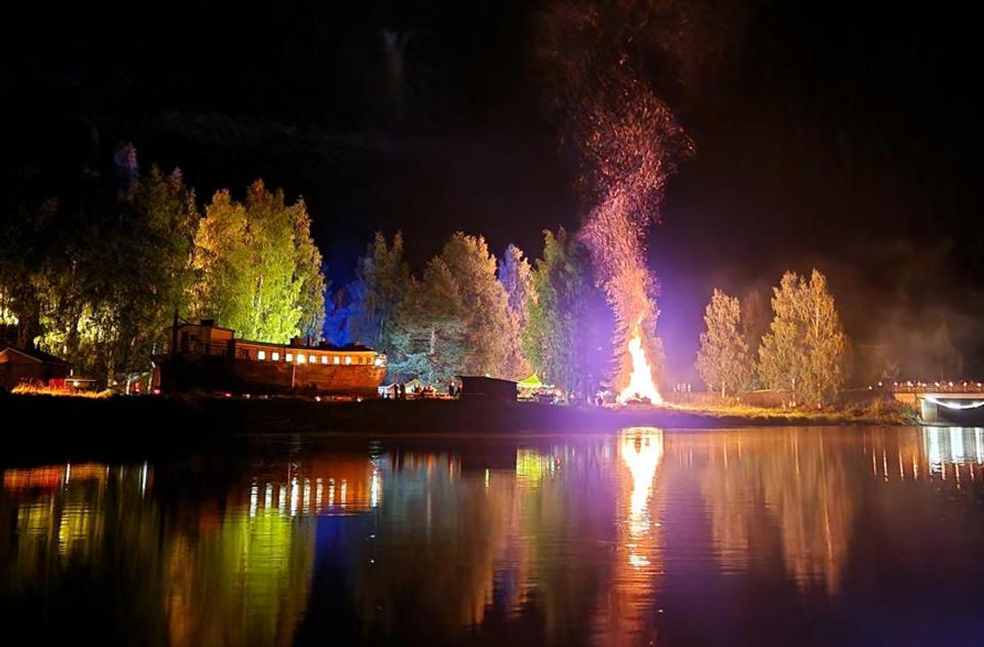 Yökuva Möhköstä vesistön äärellä järjestetystä tapahtumasta. Keskellä hehkuu suuri, korkea kokko tai roihuava nuotio, jonka liekit valaisevat taivaan. Kokko heijastuu kirkkaana tyyneen veteen etualalla. Rantaviivan puita on valaistu erivärisillä jouluvaloilla, luoden keltaisia, vihreitä ja violetteja sävyjä pimeään. Taustalla näkyy rakennuksen siluetti.