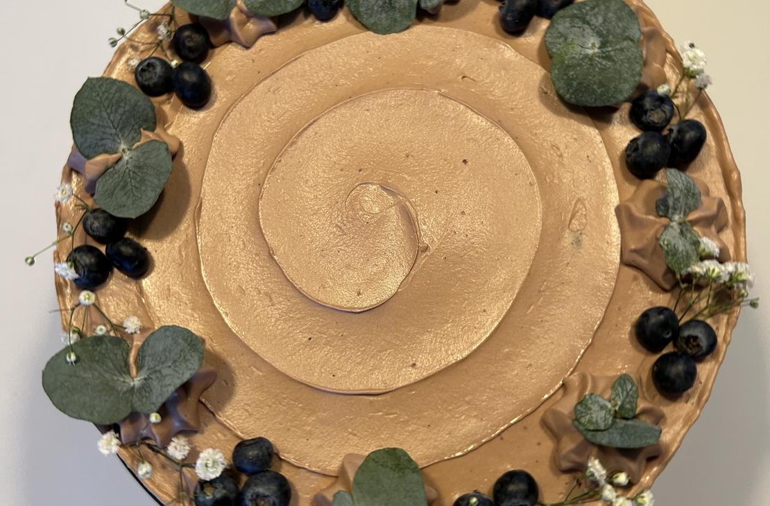 Yläviistosta otettu kuva vaaleanruskealla kuorrutteella päällystetystä kakusta, joka on koristeltu reunoilta mustikoilla, pienillä valkoisilla kukilla ja eukalyptuksen lehdillä.