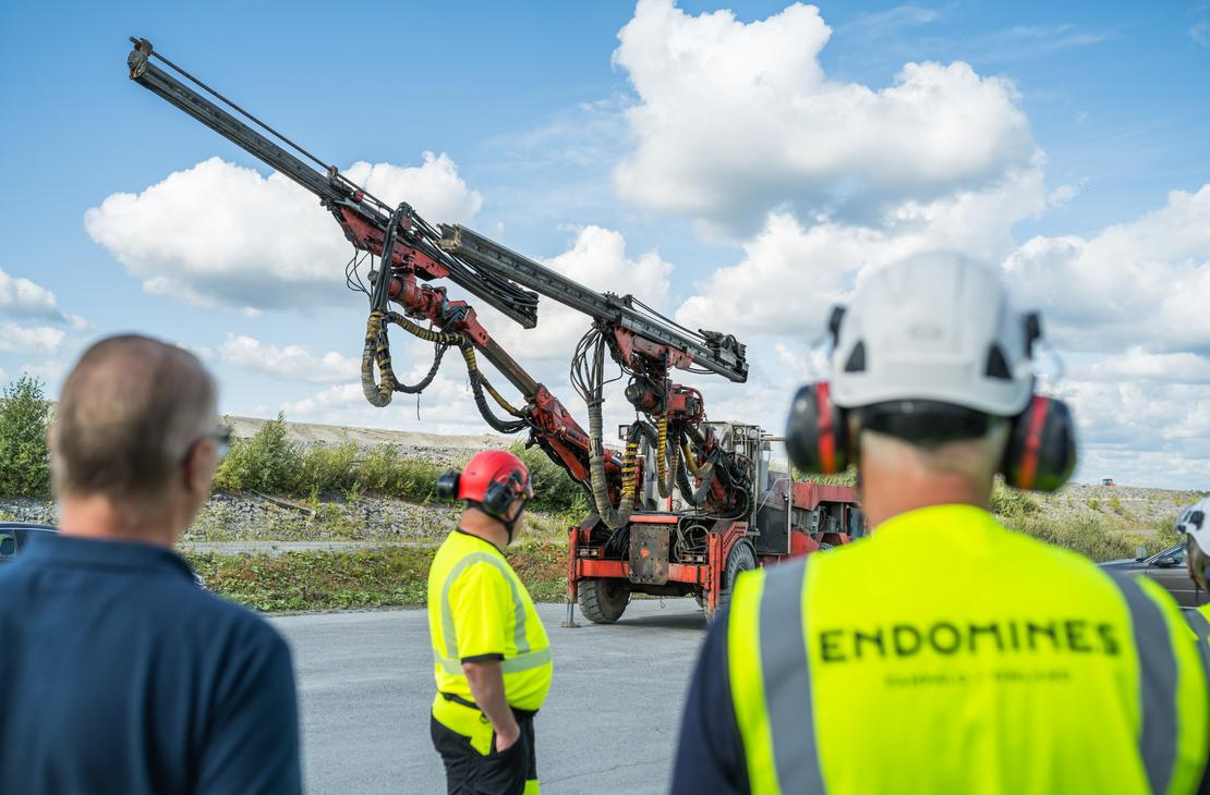 Kolme henkilöä seisoo selin kameraan ja katsoo suurta punaista kaivosporauslaitetta, jonka pitkä puomi on suunnattu viistosti kohti taivasta. Etualalla oikealla olevalla henkilöllä on päässään valkoinen suojakypärä ja yllään keltainen huomioliivi, jonka selässä lukee "ENDOMINES". Tapahtuma sijoittuu sorakentälle aurinkoisena päivänä, ja taustalla on sininen taivas valkoisilla pilvillä.