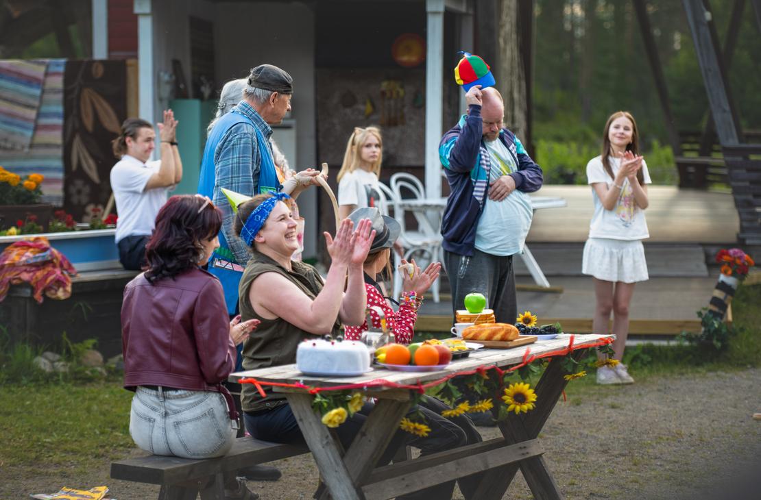 M&ouml;hk&ouml;n kes&auml;teatterin Peitetarina-n&auml;ytelm&auml;n kohtaus, jossa useita n&auml;yttelij&ouml;it&auml; on kokoontunut ulkon&auml;ytt&auml;m&ouml;ll&auml; herkuilla ja auringonkukilla koristellun pitk&auml;n puup&ouml;yd&auml;n &auml;&auml;reen. Vasemmalla etualalla sinip&auml;&auml;hineinen nainen nauraa ja taputtaa k&auml;si&auml;&auml;n viininpunaiseen nahkatakkiin pukeutuneen naisen vieress&auml;. Taustalla mies kohottaa v&auml;rik&auml;st&auml; potkurilippist&auml;&auml;n muiden n&auml;yttelij&ouml;iden taputtaessa ja seuratessa tilannetta.