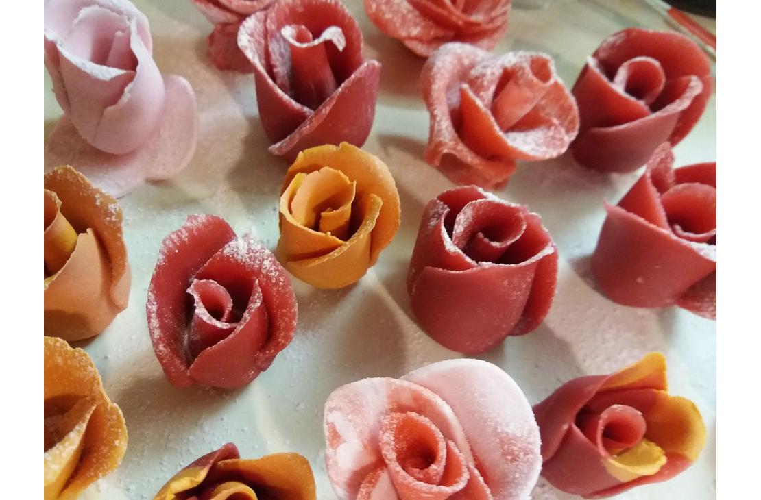 Lähikuva useista syötävistä ruusunmuotoisista koristeista tai makeisista, jotka on tehty sokerimassasta tai marsipaanista, mahdollisesti Kotipari Lämmin Halaus Oy:n tuotteita. Ruusut ovat eri sävyisiä, mukaan lukien vaaleanpunainen, koralli ja oranssi, ja ne on aseteltu vaalealle pinnalle.