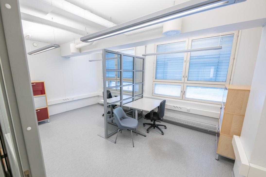 Kuva esittää Liikekulman Toimistohotellin sisätiloja. Käytävä on valoisa ja moderni, jossa on vaalea lattia ja valkoiset seinät. Oikealla on pitkä käytävä, jossa on oviaukkoja ja työpöytiä. Vasemmalla on pyöreitä mustia pöytiä ja tuoleja. Keskellä näkyy kirkkaan limevihreä ovi, jossa on logo, ja taaempana oranssin ja keltaisen sävyisiä ovia, jotka viittaavat erilaisiin toimistoihin.