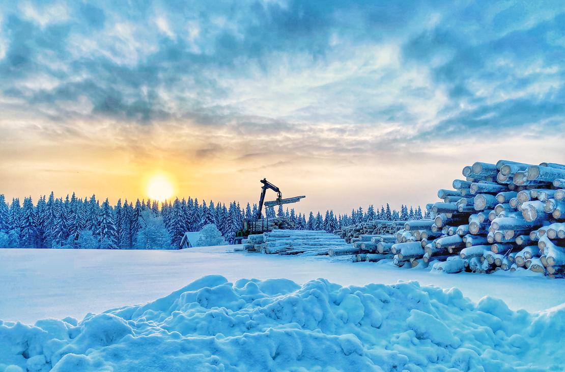 Talvisessa, laskevan auringon pakkasmaisemassa oleva John Deere ajokone, joka purkaa puita kuormasta varastopaikkaan. 