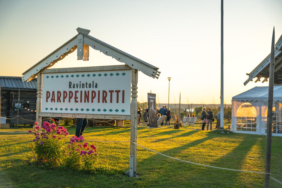 Lähikuva puisesta "Ravintola Parppeinpirtti" -kyltistä ilta-auringon paisteessa. Etualalla on kukkia, ja taustalla näkyy ihmisiä ravintolan pihapiirissä.