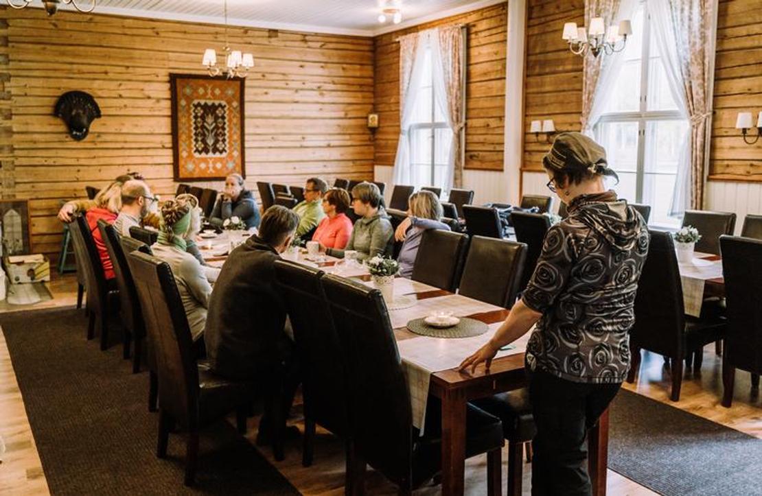 Sisäkuva Mantan Majatalon ruokasalista tai juhlatilasta. Tila on sisustettu perinteiseen tyyliin, jossa on vaaleat puupaneeliseinät ja valkoiset verhot ikkunoissa. Keskellä tilaa on pitkä pöytä, jonka ääressä on mustat tuolit ja istuu ryhmä ihmisiä. Oikealla etualalla seisoo yksi henkilö selin kameraan. Takaseinällä on kaksi koriste-esinettä, joista toinen on tumma taulu ja toinen mahdollisesti eläimen pää.