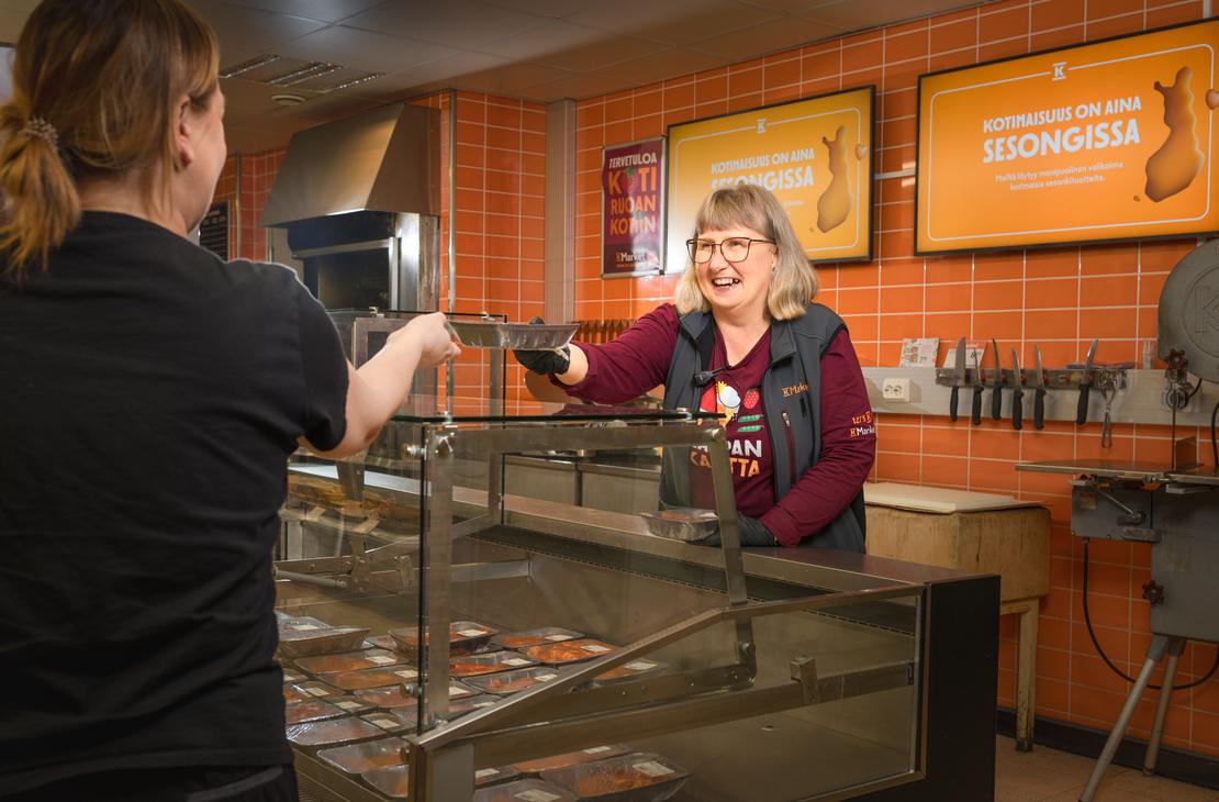 Hymyilevä K-Market Ilomantsin työntekijä ojentaa palvelutiskillä tuotetta metallivuuassa asiakkaalle lasisen suojatiskinsä yli. Taustalla on oranssi laatoitettu seinä, jossa on kyltit tekstillä ”KOTIMAISUUS ON AINA SESONGISSA”. Tiskin sisällä on esillä pakattuja tuotteita.