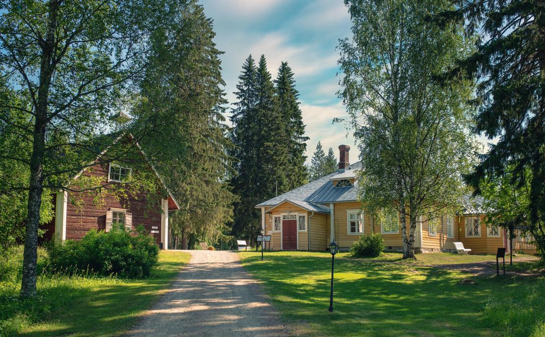 Kuva Möhkön ruukkimuseon alueelta esittää historiallista pihapiiriä. Oikealla on pitkä, matala, keltainen puutalo punaisella ovella ja harmaalla katolla. Vasemmalla näkyy korkeampi, punamultainen ulkorakennus tai aitta. Rakennusten välissä kulkee hiekkainen tie, ja niitä ympäröi vanhoja koivuja ja havupuita aurinkoisessa kesäympäristössä.