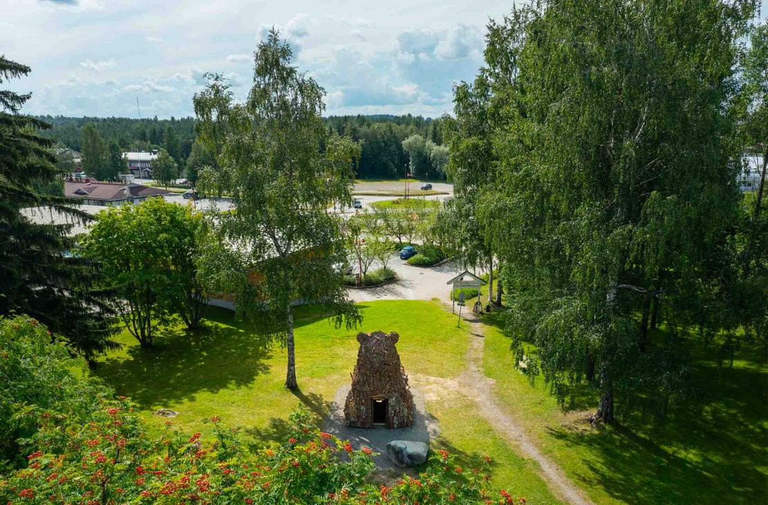 Ilmakuva suuresta, puusta tehdystä karhupatsaasta, ILOKARHU -nimisestä veistoksesta Ilomantsista. Karhu on koottu useista pienemmistä puupaloista ja laudoista. Kuvassa näkyy selvästi karhun karkea pintakerros. Taustalla on Ilomantsin tori.