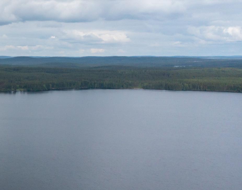 Laaja panoraamakuva, joka on otettu korkealta. Se esittää tyyntä, tummansinistä järven pintaa, joka täyttää suurimman osan kuvasta. Järven takana horisontissa näkyy tiheä, tummanvihreä metsä ja taustalla kaukaisia metsäisiä kukkuloita. Taivas on pilvinen ja vaaleanharmaa.