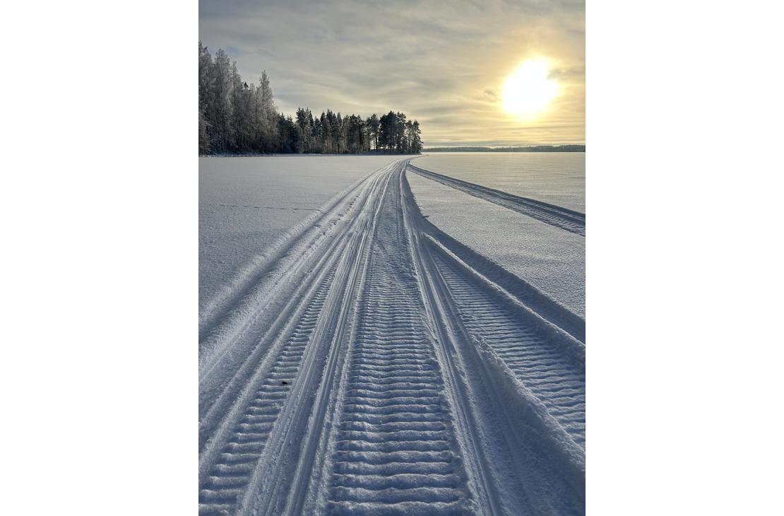 Perspektiivikuva Moottorikelkka urasta, joka kulkee lumisella järven jäällä kohti matalalla paistavaa aurinkoa ja kaukana häämöttävää metsänreunaa.