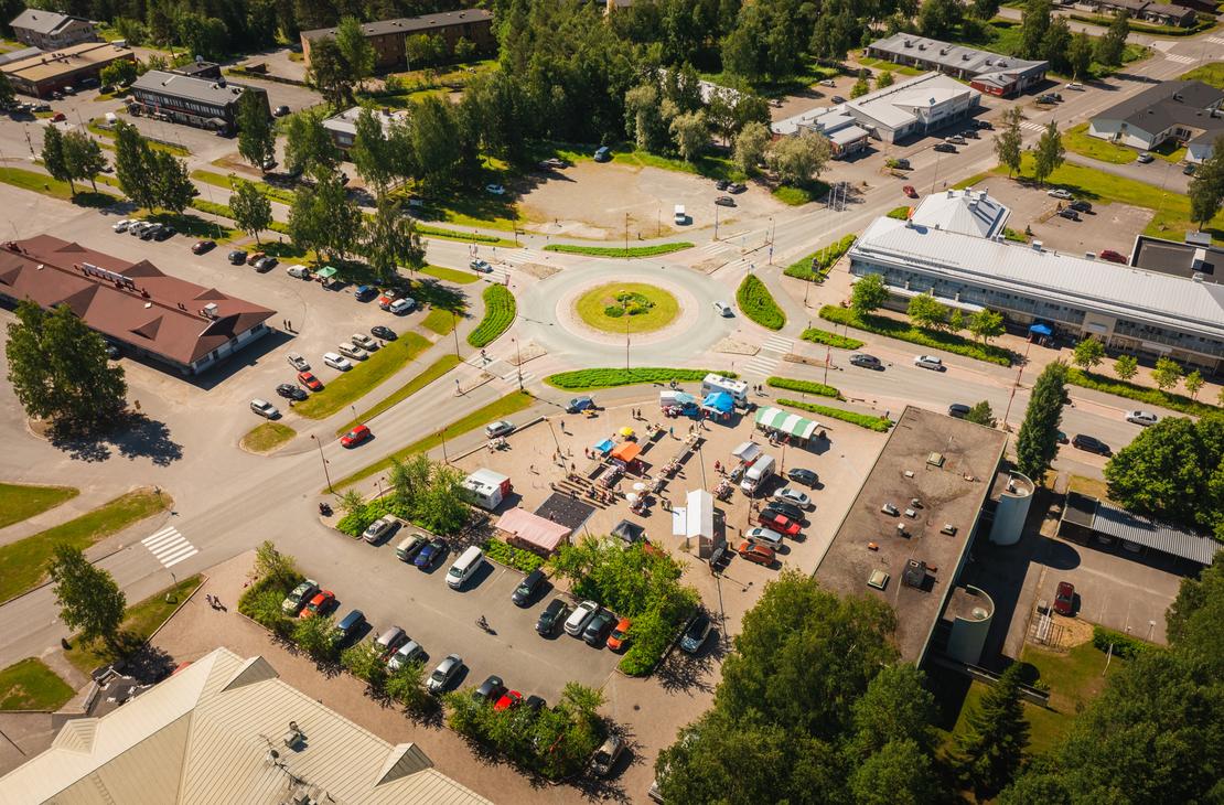 Korkealta otettu ilmakuva Ilomantsin keskustan liikenneympyrästä ja sen vieressä sijaitsevasta torialueesta, jossa on värikkäitä myyntitelttoja, ihmisiä ja ympärillä runsaasti pysäköityjä autoja aurinkoisena kesäpäivänä.